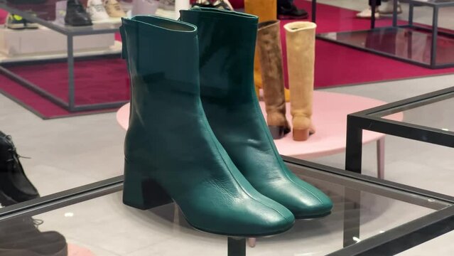 Women's dark green leather high-heels boots stand in a shoe store window. View through the glass
