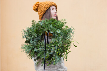 Young woman in ginger hat hiding behind a Christmas wreath. Florist holding handmade chaplet with pine and fir branches.