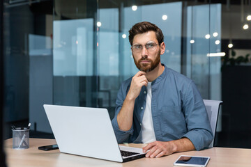 Fototapeta na wymiar .Serious pensive businessman in shirt thinking about decision sitting at table in modern office, man with beard is using laptop at work.