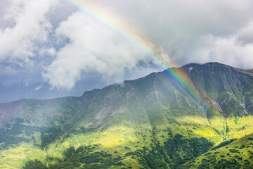 A rainbow shines through atmospheric light, lush green mountainsides in the background, Hatcher Pass, South-central Alaska; Palmer, Alaska, United States of America