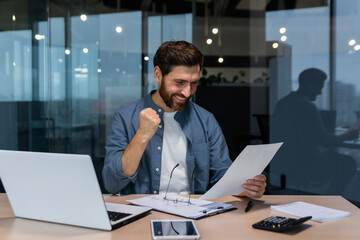 Successful businessman in casual shirt doing paperwork, boss with beard and glasses sitting at desk at workplace using laptop working with documents, holding hand up celebrating victory and triumph.