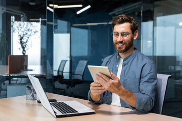 Successful businessman in a shirt uses a tablet computer, a man sits at a desk smiling and happy, a...