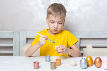 A little blond boy paints eggs for the Easter holiday at home. The child has fun and celebrates the holiday.