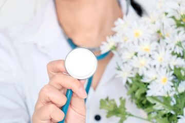 Obraz na płótnie Canvas The doctor is young, an Asian girl with black hair in medical white clothes with a stethoscope around her neck. Holds a bouquet of chamomile flowers in her hands. Light background. Medic day 