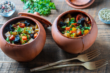 Stewed potato, carrot, onion, tomato and prunes in a clay pots with lid on wooden background,...