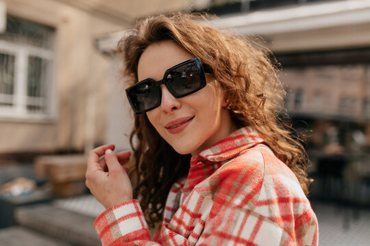 Outdoor close up portrait of smiling stylish adorable lady with curly hair wearing black sunglasses and bright shirt posing at camera in sunlight with adorable smile