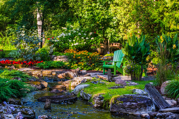A tranquil stream flows through a garden area with a green adirondack chair; Hudson, Quebec, Canada