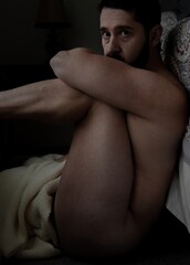 attractive male model sitting naked on floor against bed holding head with muscular arms showing side of bare nude skin with dark background vertical