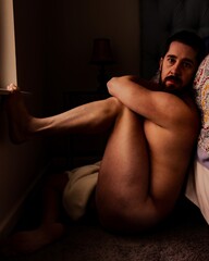 Intimate attractive nude male model sitting on floor against bed holding head with muscular arms showing side of bare naked skin with dark background vertical