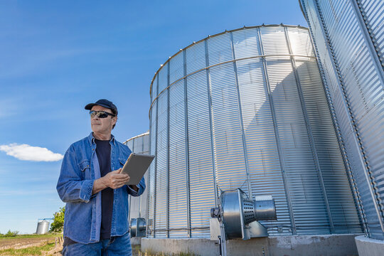 A farmer stands beside grain storage bins using a tablet; Alberta, Canada