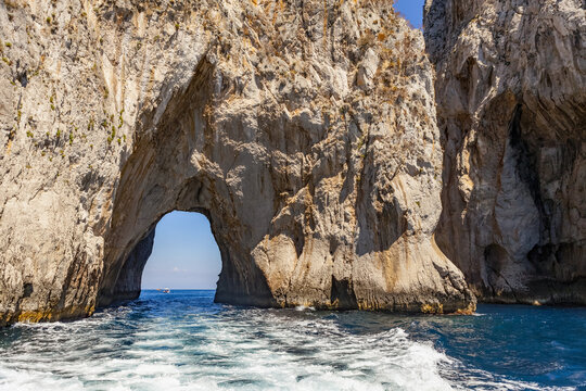 Natural arch in the limestone rock formations of the Faraglioni Rocks with a view of a boat, Island of Capri on the Tyrrhenian Sea, Mediterranean; Capri, italy