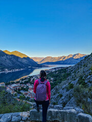 Naklejka na ściany i meble Woman with panoramic view from the Ladder of Kotor on the Kotor bay during sunrise, Adriatic Mediterranean Sea, Montenegro, Balkan Peninsula, Europe. Winding fjord water reflection along coastal towns