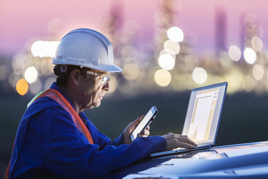 Man working on a laptop and smart phone with an oil refinery in the background; Alberta, Canada