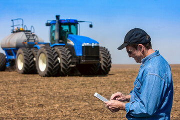 Farmer using a tablet while standing on a farm field and a tractor and equipment seeds the field; Alberta, Canada