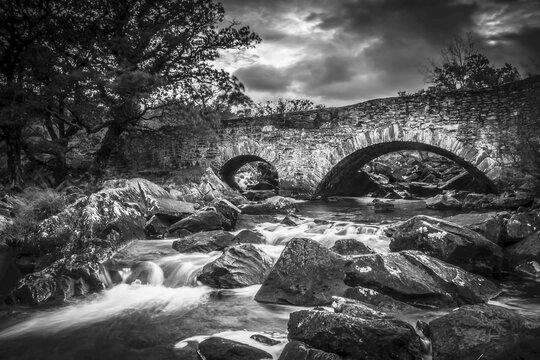 Black and white image of an arched stone bridge over a flowing river; County Kerry, Ireland