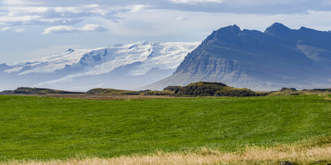Landscape in Southeastern Iceland with rugged mountains and grass field, and snow-covered mountains in the distance; Hornafjorour, Eastern Region, Iceland