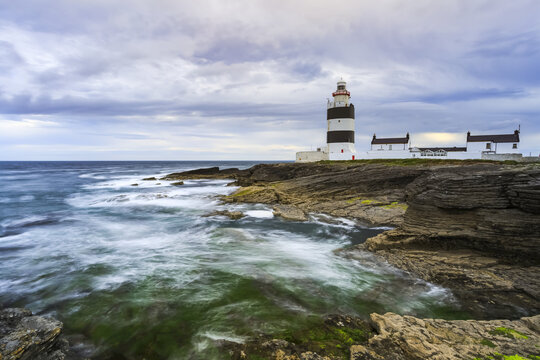 Hook Head lighthouse, Black and white lighthouse on rocky shore on a cloudy summer day., long exposure; County Waterford, Ireland