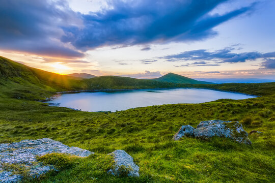 Sun setting over Lough Muskery in the Galty Mountains in summer with large boulders in the foreground, Galty Mountains; County Limerick, Ireland
