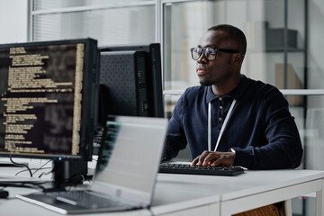 African American young developer in eyeglasses concentrating on his online work on computer sitting at workplace - obrazy, fototapety, plakaty