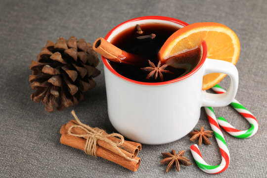White mug with mulled wine, cinnamon stick, clove, slice of orange and anise inside of it, candy canes, cone, bunch of cinnamon sticks, gray fabric