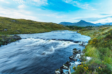 Small rapids on a river flowing through Connemara with a mountain in the background on a blue sky day; Connemara, County Galway, Ireland
