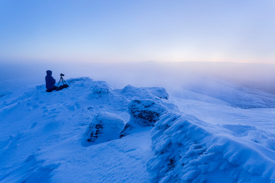 Man kneels beside his tripod and camera on a snow-covered mountain in the fog, Galty Mountains; County Tipperary, Ireland