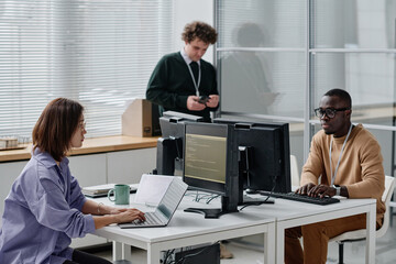 Group of young developers writing codes for new computer program sitting at table with computers in...