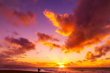 Silhouette of a surfer standing on Kelki Beach at the water's edge with dramatic glowing clouds above at sunset; Oahu, Hawaii, United States of America