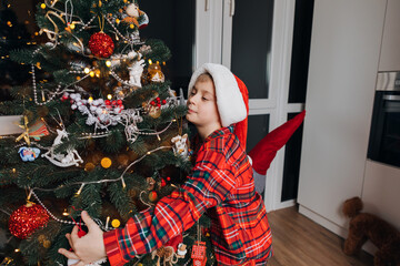 A boy in red pajamas and Santa hat hugging a Christmas tree at home. Christmas and family. Front view