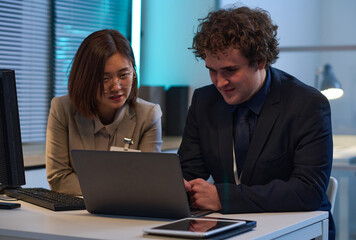 Young colleagues discussing computer program on laptop while working in team in office till late evening