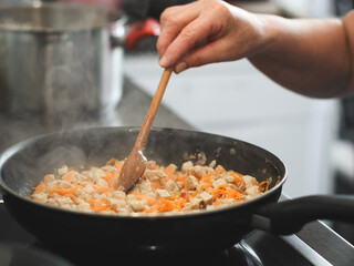 The hand of a senior woman holds a wooden spoon and stirs meat with carrots that are fried