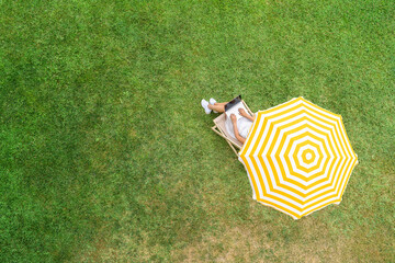 Woman in a white dress sitting on deck chair under yellow umbrella with laptop on the green grass...