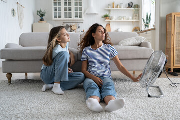 Loving carefree mother and teenage girl sits in relaxed position on floor in living room near fan....