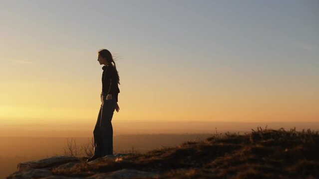 A young inspired woman walks to the edge of a cliff and raises her arms to the sides while standing on top of a mountain against a yellow sky. Watching the sunset with a beautiful view