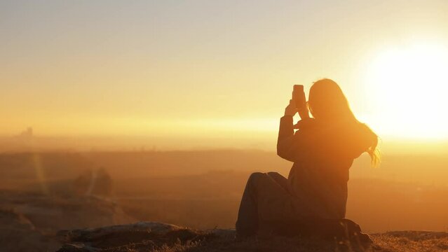  Silhouette of happy smiling woman girl against yellow sky at sunset sitting on top of mountain with incredible view and taking photo on phone camera, traveler, outdoor walk