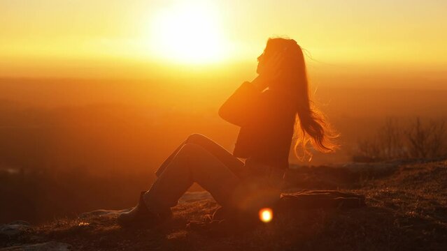 Young long-haired woman in modern wireless headphones relaxing on top of mountain with incredible view at sunset listening to music. Happy young girl puts on headphones and enjoys quality sound. 