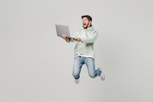 Full body young caucasian happy surprised excited amazed IT man wear mint hoody jump high hold use work on laptop pc computer isolated on plain solid white background studio. People lifestyle concept.