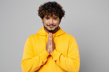Obraz premium Young asking Indian man 20s he wearing casual yellow hoody holding hands folded in prayer gesture, begging about something isolated on plain grey background studio portrait. People lifestyle portrait.