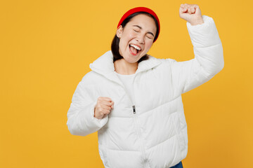 Young overjoyed woman of Asian ethnicity wear white padded windbreaker jacket red hat do winner gesture celebrate clenching fists isolated on plain yellow background studio People lifestyle concept.