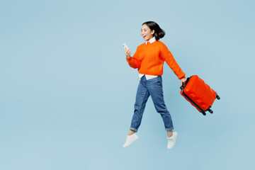 Full body young traveler woman of Asian ethnicity wear orange sweater hold valise mobile phone...