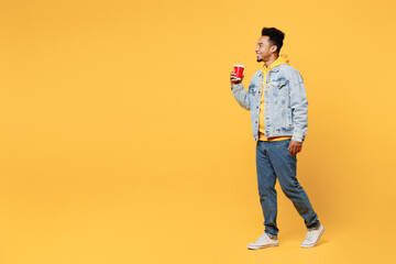 Fototapeta premium Full body side view young man of African American ethnicity wear denim jacket hoody hold takeaway delivery craft paper brown cup coffee to go walk isolated on plain yellow background studio portrait.