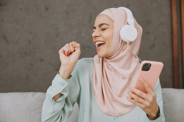 Young muslim woman wears hijab casual clothes headphones listen music use mobile phone sing sit on...