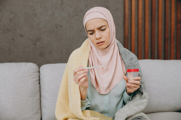 Young muslim woman wear hijab casual clothes plaid check thermometer drink pill sits on sofa stay at home flat rest relax spend free spare time in living room People middle eastern uae islam concept.