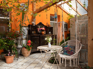 Interior of veranda. Cozy space in patio. A lot of plants. Orange wall. Wooden furniture. Hanging wicker white chair.