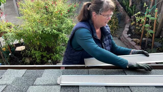 4K HD video of an older caucasian woman wearing basic gardening gloves installing gutter guards under shingles of roof over rain gutters on house. view from above with backyard in background.
