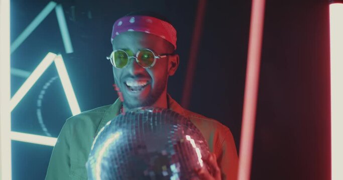 Slow motion of good-looking African American man dancing with disco ball on neon lamps background. Entertainment and clubbing concept.