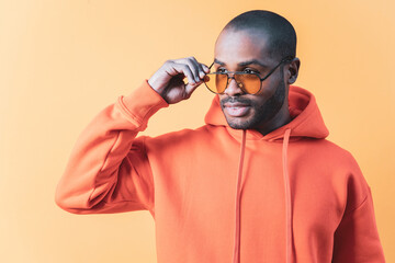 The portrait of a Colombian man in an orange sweatshirt posing with sunglasses on an orange background, the man looks over the glasses. African American fashion and style concept.