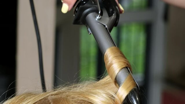Anonymous stylist does perm using stainless curling iron in beauty salon. Hairdresser makes modern hairstyle for blonde client close backside view