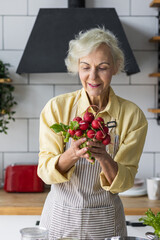 Attractive senior woman with grey hair cooking healthy food on her kitchen at home. Mature female preparing fresh vegetarian salad with organic ingredients from the market: tomato, cucumber, pepper