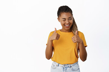 Portrait of beautiful young black woman shows thumbs up, winks and smiles pleased, approves smth good, recommends store or app, white background